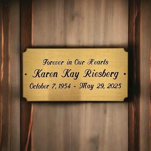 Może przedstawiać: Złota tablica z napisem "Forever in Our Hearts" i imieniem "Karen Kay Riesberg". Zawiera również daty "October 7, 1954 - May 29, 2025". Tablica jest zamontowana na drewnianej powierzchni.