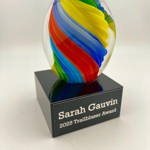 May include: A clear glass award with a colorful, swirled design in red, blue, yellow, and green, set on a black base. The base is engraved with "Sarah Gauvin" and "2025 Trailblazer Award."