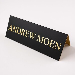 May include: A black triangular desk nameplate featuring the name ANDREW MOEN printed in gold capital letters. The sign is folded into a tent shape with a matte black exterior finish and visible gold edges on the side panel of the structure.