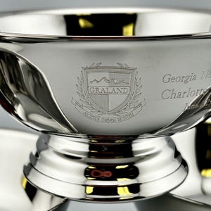 May include: A close-up of a silver trophy bowl with a decorative base. The bowl has an engraved crest with the word "GRALAND" and the Latin phrase "ASCENDE OMNEM MONTEM". The name "Georgia Ne" and "Charlotte" are also engraved.