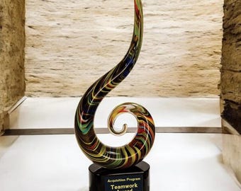 May include: A colorful glass sculpture with a swirling, spiral design. The sculpture is mounted on a black base with a plaque that reads "Acquisition Program Teamwork Award."