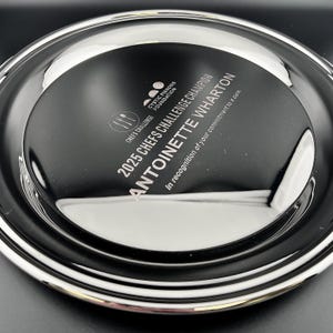 May include: A silver-colored plate with a black center. The center features text that reads "2025 CHEFS CHALLENGE CHAMPION ANTOINETTE WHARTON" and "In recognition of your commitment to a cure."