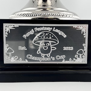Pu&ograve; includere: Un trofeo nero con la parte superiore argentata. La parte anteriore del trofeo presenta una placca nera con le parole "Fungi Fantasy League Champion's Cup" e un disegno di fungo dei cartoni animati. Viene visualizzato anche l'anno 2023.