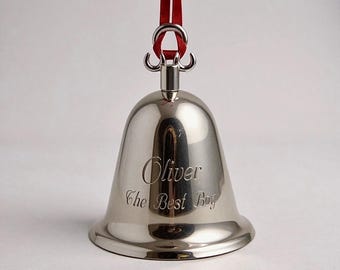 Pode incluir: Um ornamento de sino em metal prateado polido com uma fita vermelha no topo. O sino apresenta a gravação do texto "Oliver" na linha superior e "The Best Boy" na linha inferior, utilizando uma fonte cursiva.