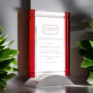 May include: A clear glass retirement award with red accents and a silver base. The award features the text "RETIREMENT AWARD" and "JEFFREY PLANTER". The award is displayed on a gray surface with green foliage in the background.