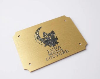 May include: A rectangular brushed gold metal plaque with rounded corners and four small circular holes at the edges. The center features a black graphic of a crescent moon and crystals with the text LUNA MUNA COUTURE printed below.