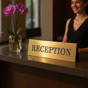 May include: A gold-colored metal sign with the word "RECEPTION" in black lettering sits on a dark countertop. A vase of purple orchids is to the left. A person in a black top is visible in the background.