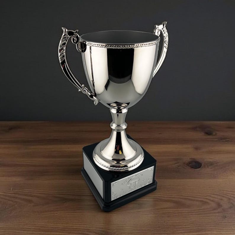 Pu&ograve; includere: Un trofeo d'argento lucido con manici decorati e una base nera. Il trofeo ha una coppa ampia e un bordo dettagliato. La base ha una placca d'argento. Posizionato su una superficie di legno.