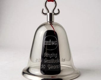 Pode incluir: Um sino de metal prateado polido com um laço de fio vermelho no topo. O sino apresenta uma incrustação retangular preta gravada com o texto "miec" dentro de um círculo, seguido pelas palavras "Ringing with Gratitude for 50 Years!"
