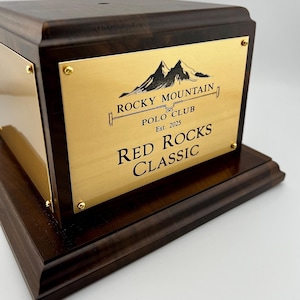 Könnte beinhalten: Dunkle Holzbox mit goldener Plakette mit der Aufschrift "Rocky Mountain Polo Club" und "Red Rocks Classic". Die Plakette zeigt auch eine Berg-Grafik und das Jahr "Est. 2025". Goldene Schrauben befestigen die Plakette an der Box.