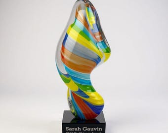 May include: A colorful glass sculpture with a swirling design, featuring bands of blue, yellow, orange, and green. The sculpture sits on a black base with the text "Sarah Gauvin 2086 Trailblazer Award."