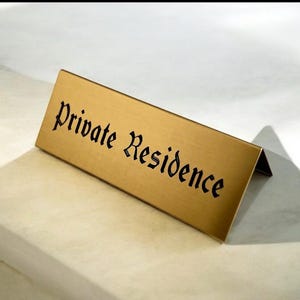 May include: A gold-colored metal sign with the words "Private Residence" in black, stylized lettering. The sign is angled, with a folded base for standing. The background is a light-colored surface.