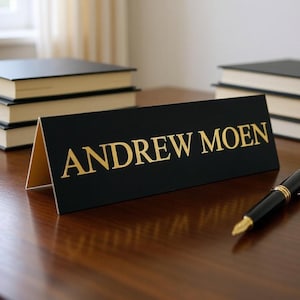 May include: A black triangular desk nameplate with the name ANDREW MOEN printed in gold capital letters. The nameplate is positioned on a dark wooden surface next to a black fountain pen, with stacks of books visible in the background.