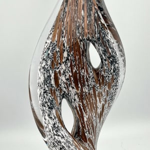 Puede incluir: Una escultura decorativa de vidrio con un exterior claro y curvado y un interior con un patrón de motas marrones, blancas y negras. La escultura tiene dos aberturas ovaladas y una forma orgánica y fluida.