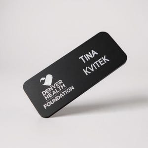 May include: A black rectangular name tag with rounded corners. The name tag features the name "TINA KVITEK" in white text. The Denver Health Foundation logo is also displayed in white.
