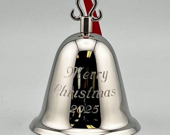 Pode incluir: Um ornamento de sino em tom prateado polido com um laço de metal decorativo no topo e uma fita vermelha. A parte frontal do sino apresenta a inscrição gravada Merry Christmas 2025 em uma fonte de estilo cursivo clássico.