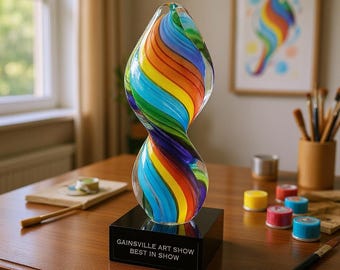May include: A colorful glass sculpture with a twisted, hourglass shape, featuring vibrant rainbow stripes. It sits on a black base with the text "GAINSVILLE ART SHOW BEST IN SHOW". The artwork is displayed on a wooden table.