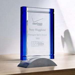 May include: A clear glass award with blue accents and a gray base. The award features the Verizon logo, the name "Suzy Hagglund", and the "Flanagan Spirit Award" text. The year "2005" is also visible.
