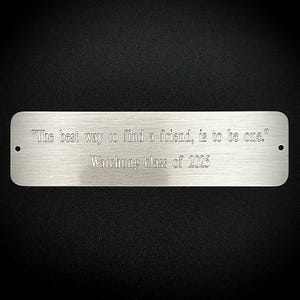 Puede incluir: Placa rectangular de metal cepillado con esquinas redondeadas. El texto grabado dice: "La mejor manera de encontrar un amigo es ser uno." y "Watchung class of 2025." Dos pequeños agujeros perforados en cada extremo.