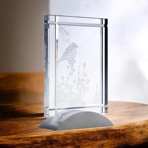 May include: A clear glass rectangular decorative piece with a bird and floral design etched inside. The piece is mounted on a gray, curved base and sits on a wooden surface. The glass is thick and has a polished finish.