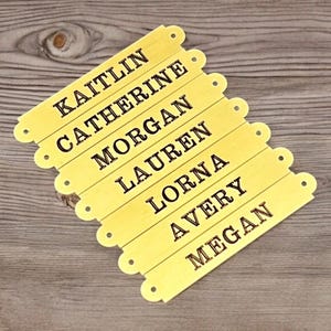 Op de afbeelding: Een set van acht gele houten naamlabels met zwarte tekst. De namen zijn Kaitlin, Catherine, Morgan, Lauren, Lorna, Avery en Megan. Elk label heeft een decoratieve rand en kleine gaatjes.