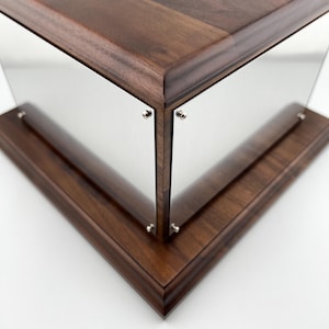 Könnte beinhalten: Eine dunkle, braune Holzvitrine mit klaren, reflektierenden Paneelen. Die Paneele werden von kleinen, silberfarbenen Befestigungselementen gehalten. Das Design ist eckig und modern, mit Fokus auf klaren Linien.