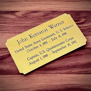 May include: A gold-colored plaque with rounded corners, featuring engraved text. The text reads "John Kemanai Warner" and details his service in the U.S. Army, including dates of service as Lieutenant and Captain. The plaque rests on a wooden surface.