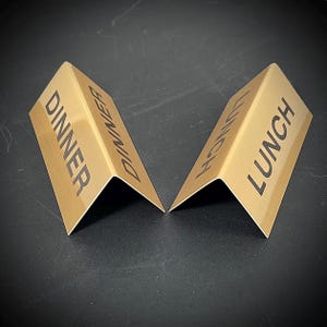 May include: Two gold-colored, angled table signs. One sign reads "DINNER" in black capital letters, and the other reads "LUNCH." The signs are made of a metallic material and are designed to stand upright.