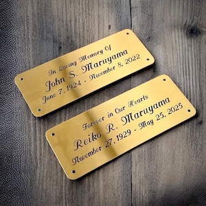 Könnte beinhalten: Zwei rechteckige, goldfarbene Gedenktafeln mit graviertem Text. Die obere Tafel lautet: "In Loving Memory Of John S. Maruyama June 7, 1924 - November 8, 2022." Die untere Tafel lautet: "Forever in Our Hearts Reiko R. Maruyama November 27, 1929 - May 25, 2025."
