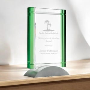 May include: A clear glass award with green edges and a gray base. The award features the text "Pacific Palms Golf Club Distinguished Member Award" and the name "James Patterson". The award is displayed on a wooden surface.