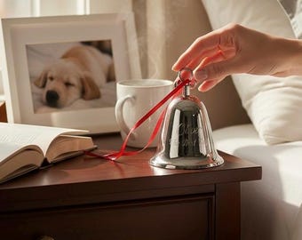Pode incluir: Um sino de metal prateado polido com uma fita vermelha presa à alça superior. O sino apresenta uma inscrição gravada que diz Oliver e Our Best Wish. O sino está posicionado ao lado de uma caneca de cerâmica branca e um livro aberto.