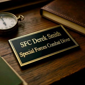 May include: A rectangular black plaque with a gold border and gold engraved text that reads SFC Derek Smith Special Forces Combat Diver. The plaque is positioned near a round metallic compass and a brown leather-bound book.