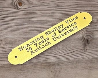 Può includere: Targa dorata con estremità arrotondate, con il testo inciso: "Honoring Shelley Viles 34 Years of Service Antioch University". La targa ha due piccoli fori per il montaggio e poggia su una superficie di legno.