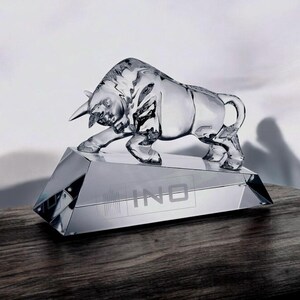 May include: A clear glass sculpture of a bull in a charging stance, mounted on a faceted base. The base has the word "INO" etched on it. The sculpture is displayed on a dark wooden surface.