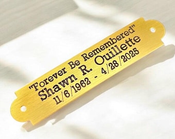 May include: A gold-colored metal plaque with rounded ends and two holes. The plaque has the engraved text "Forever Be Remembered" above the name "Shawn R. Ouillette" and the dates 11/6/1962 - 4/28/2025.