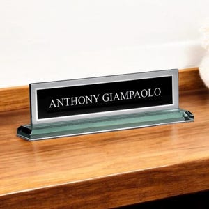 May include: A clear glass desk nameplate with the name "ANTHONY GIAMPAOLO" in white text on a black background. The nameplate sits on a wooden surface.