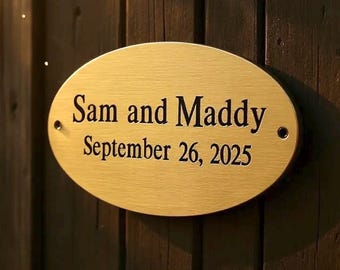 Puede incluir: Una placa ovalada de metal dorado cepillado con el texto negro grabado "Sam and Maddy" en la línea superior y "September 26, 2025" en la línea inferior. La placa incluye dos pequeños orificios de montaje circulares en los lados izquierdo y derecho.