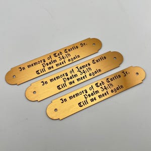 May include: Three gold-colored memorial plaques with rounded ends and a hole at each end. Each plaque has engraved text: "In memory of" followed by a name, "Psalm 34:18," and "Till we meet again."