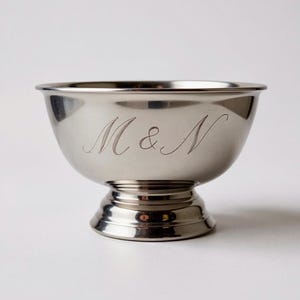 May include: A polished silver bowl with a pedestal base. The bowl has a rounded shape with a slightly flared rim. The initials "M & N" are elegantly engraved on the front of the bowl. The bowl is designed to be used for serving.