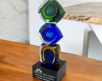 May include: A stacked glass sculpture with three colorful, square-shaped elements. The top piece is green, the middle is blue, and the bottom is yellow and blue. It sits on a black base with text that reads "CREATIVE ARTS GROUP".