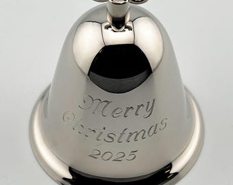 Pode incluir: Um ornamento de sino em metal prateado polido com uma superfície lisa e reflexiva. A frente do sino está gravada com o texto Merry Christmas e o ano 2025 em uma fonte cursiva. Um laço de metal está posicionado no topo do sino.