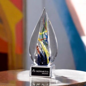 Op de afbeelding: Een heldere glazen prijs in de vorm van een druppel, met kleurrijke wervelingen van rood, geel en blauw aan de binnenkant. De basis heeft een zwart plaatje met het logo "Novartis Oncology".