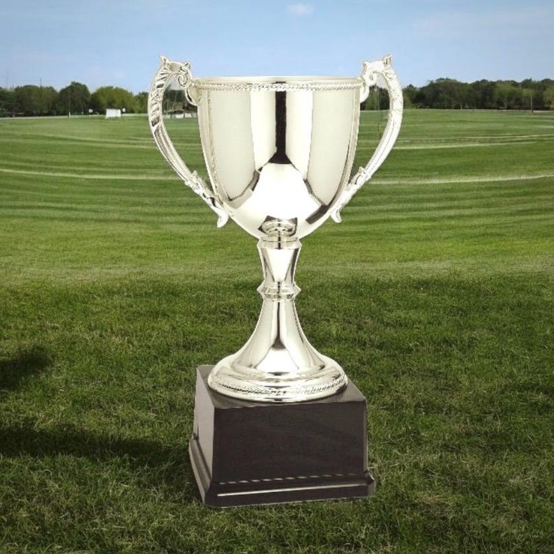 Pu&ograve; includere: Un trofeo d'argento lucido con manici e dettagli ornati, posto su una base quadrata nera. Il trofeo &egrave; esposto su un campo erboso verde sotto un cielo blu, suggerendo un premio o un riconoscimento.