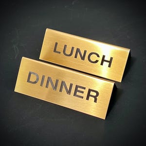 Puede incluir: Dos letreros rectangulares de metal dorado. Un letrero dice "LUNCH" en letras mayúsculas negras y en negrita. El otro letrero dice "DINNER" en la misma fuente. Los letreros están ligeramente inclinados.
