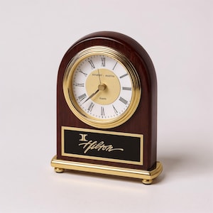 Peut inclure: Une horloge de bureau en bois avec une lunette et une base dorées. Le cadran est blanc avec des chiffres romains noirs et des aiguilles dorées. L'horloge porte le logo Hilton sur une plaque noire.