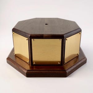 May include: A wooden, octagonal base with a dark brown finish. The base features three rectangular, gold-colored metal plates secured with small gold screws. The top has a small hole in the center.