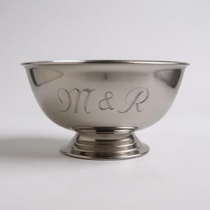 Może przedstawiać: Srebrna miska z podstawą. Misa ma zaokrąglony kształt i jest grawerowana literami "M & R" w ozdobnej czcionce. Misa jest prawdopodobnie wykonana ze stali nierdzewnej lub podobnego metalu.
