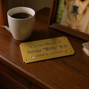 May include: A rectangular brass memorial plaque with rounded corners and four screw holes. The engraved text reads: In Loving Memory of Webster "Webby" Holt, Always in our hearts, accompanied by two small engraved paw prints on the surface.