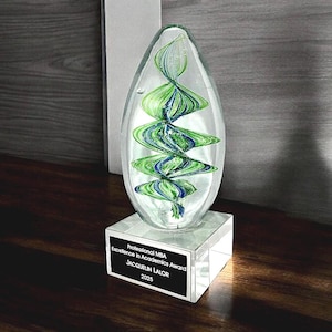 May include: A clear glass award with green and blue swirls inside, set on a clear square base. The award has a black plaque with the text "Professional MBA Excellence in Academics Award JACQUELIN LALOR 2025".