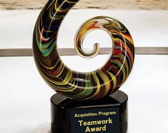 May include: A colorful, swirled glass award with a black base. The glass features streaks of red, yellow, green, and blue. The base has a plaque that reads "Acquisition Program Teamwork Award."